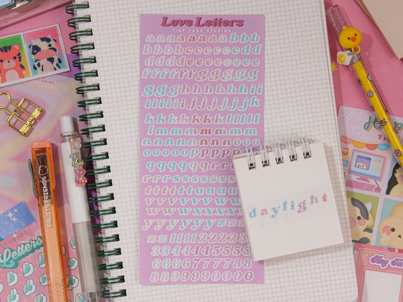 May include: A pink and white sticker sheet with the words "Love Letters" and the alphabet in a rainbow gradient. The sticker sheet is on a white notebook with a green spiral binding. A small white notebook with the word "daylight" written on it is on top of the sticker sheet.