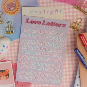 Op de afbeelding: Een vel met pastelkleurige alfabetstickers met de woorden "Love Letters" en "Tiny Taro Studios" erop gedrukt. De letters zijn in een retro, afgerond lettertype.