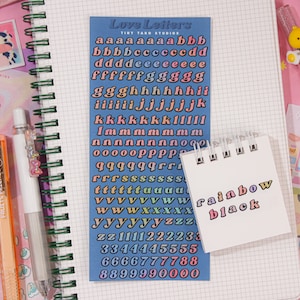 May include: A sheet of colorful alphabet stickers with a rainbow gradient. The stickers are on a blue background and include letters from A to Z and numbers from 0 to 9. The stickers are arranged in rows and columns. The sheet is on a white notebook with a green spiral binding.
