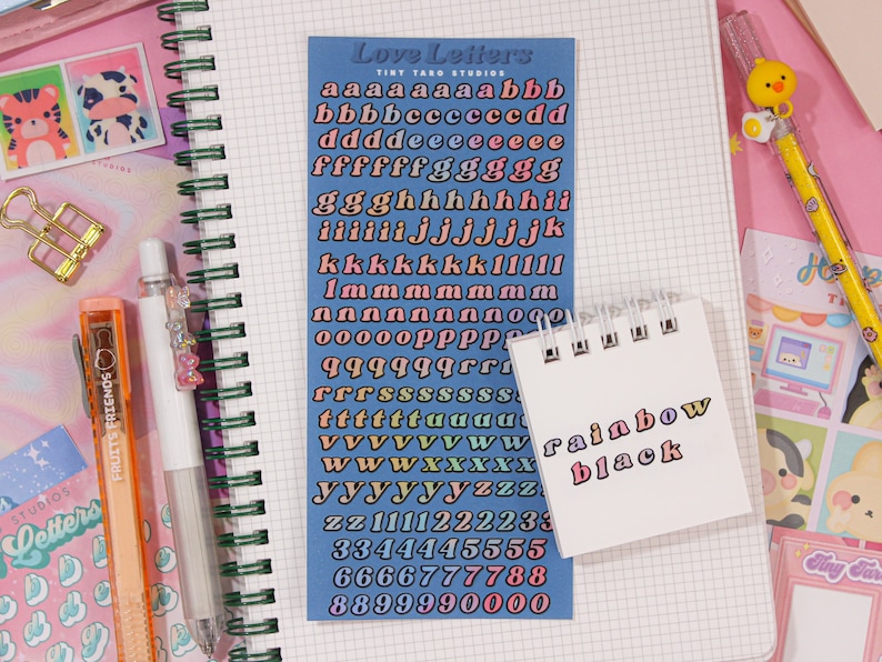 May include: A sheet of colorful alphabet stickers with a rainbow gradient. The stickers are on a blue background and include letters from A to Z and numbers from 0 to 9. The stickers are arranged in rows and columns. The sheet is on a white notebook with a green spiral binding.