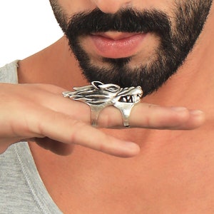 Op de afbeelding: Een zilveren ring met een wolfskopontwerp. De ring wordt gedragen aan de wijsvinger van een persoon met kort haar en een baard.