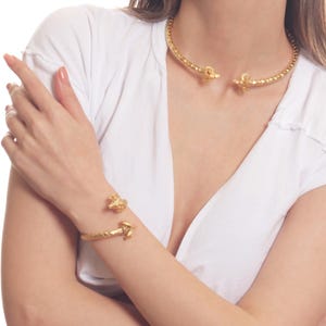 Puede incluir: Un collar gargantilla dorado y una pulsera a juego con un diseño de nudo retorcido. El collar y la pulsera son de metal y tienen una textura martillada.