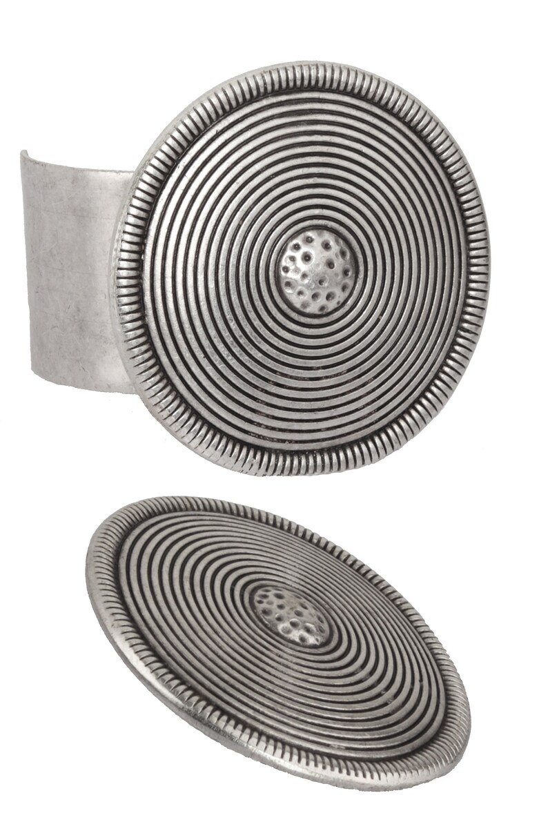May include: A silver cuff bracelet with a large, round, concentric circle design. The center of the circle has a textured, raised design.