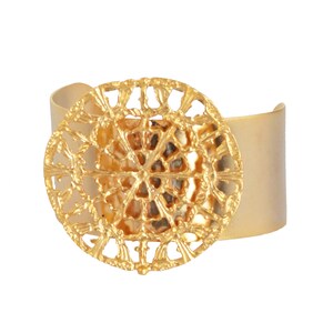 May include: A gold-toned cuff bracelet with a large, circular, openwork design in the center. The design features a repeating pattern of small, intricate shapes.