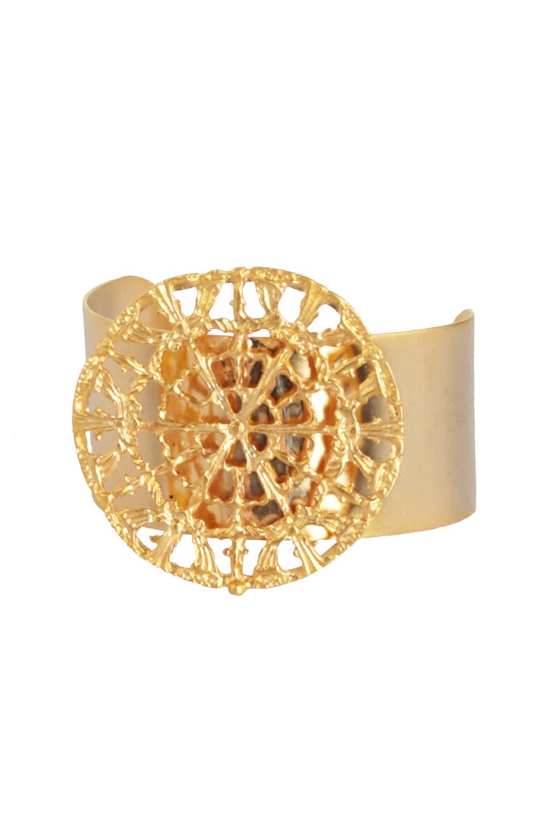 May include: A gold-toned cuff bracelet with a large, circular, openwork design in the center. The design features a repeating pattern of small, intricate shapes.