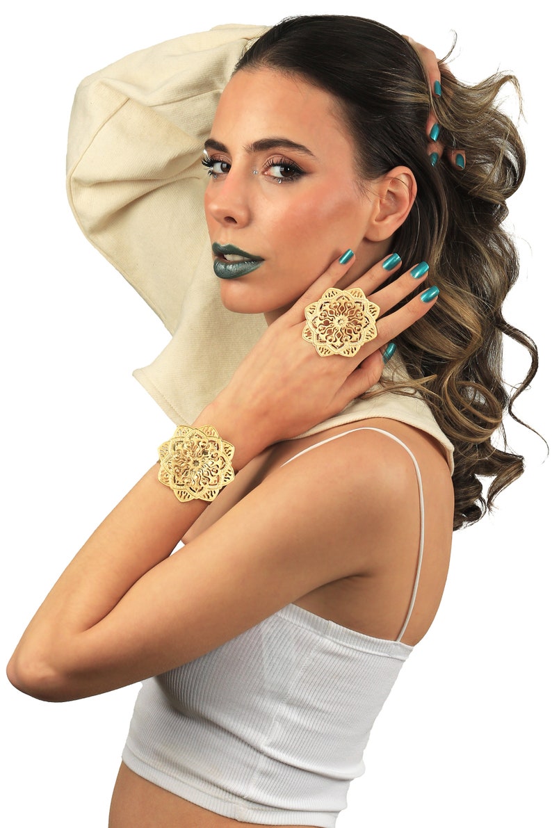 May include: A woman wearing a white top and a gold bracelet and ring with a floral design. The ring and bracelet are both intricate and detailed.