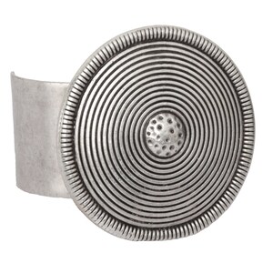 May include: A silver cuff bracelet with a large, round, concentric circle design. The center of the circle has a textured, raised dot.