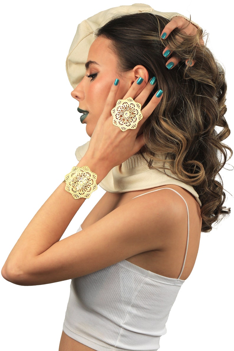 May include: A woman wearing a white top and a white sweater with a gold filigree bracelet and ring. The ring and bracelet are both in the shape of a flower.