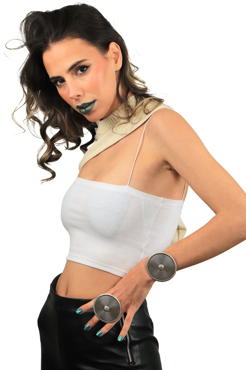 May include: A woman wearing a white crop top, black leather pants, and two silver rings with a circular design. The rings are on her wrist and finger.