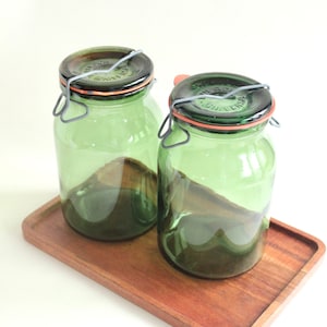 Op de afbeelding: Twee groene glazen potten met metalen sluitingen en oranje rubberen afdichtingen staan op een rechthoekige houten dienblad. De potten hebben een vintage uitstraling en zijn gedeeltelijk gevuld met een donkere substantie. De dienblad heeft een warme, roodbruine kleur.