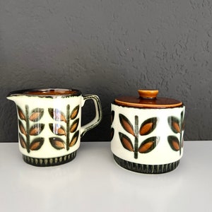 May include: A white ceramic creamer and sugar bowl set with a brown and green leaf pattern. The creamer has a handle and a spout. The sugar bowl has a lid.