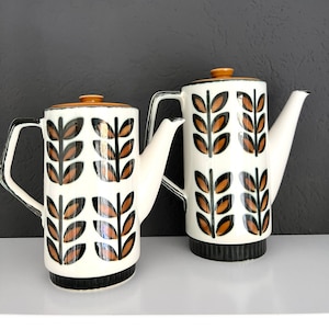 May include: Two white ceramic coffee pots with a black and orange geometric leaf pattern. The pots have a rounded body and a spout on the side. The lids are a light orange colour.