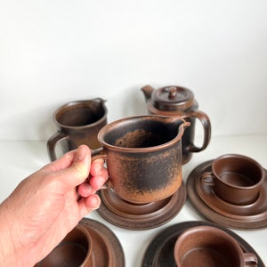 May include: A brown ceramic tea set including cups, saucers, a creamer, and a teapot. The cups and creamer have handles. The set has a textured, speckled finish. The items are arranged on a white surface.