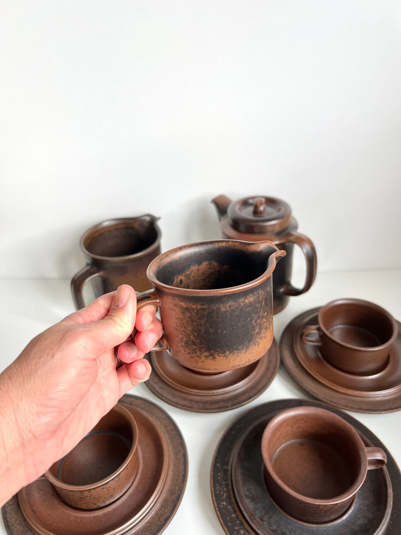 May include: A brown ceramic tea set including cups, saucers, a creamer, and a teapot. The cups and creamer have handles. The set has a textured, speckled finish. The items are arranged on a white surface.