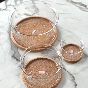 May include: Three clear glass bowls of different sizes, each on a round cork base. The bowls have a simple, modern design. The cork bases are light brown. The bowls are arranged on a white marble surface.