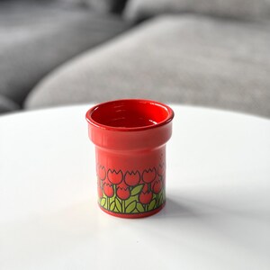May include: Red ceramic cup with a tulip pattern. The cup has a white background with red tulips and green leaves.