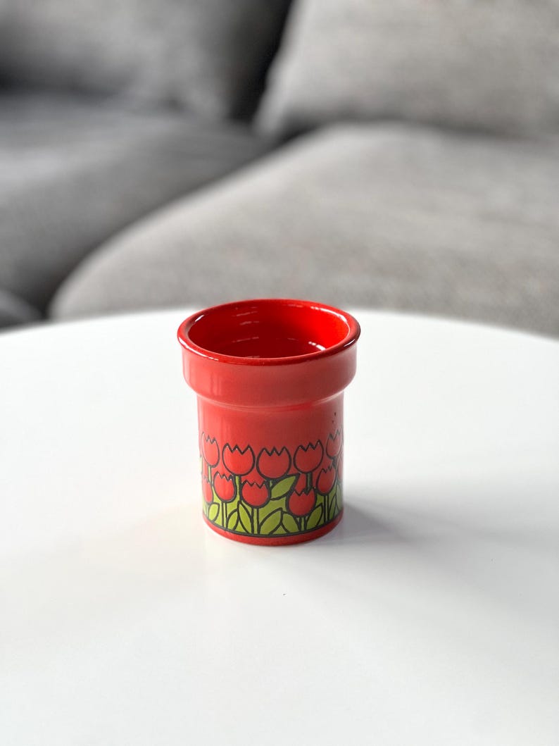 May include: Red ceramic cup with a tulip pattern. The cup has a white background with red tulips and green leaves.