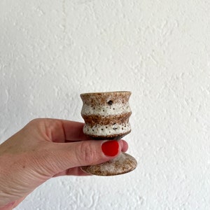 Pu&ograve; includere: Un piccolo vaso in ceramica bianca con strisce e macchie marroni. Il vaso ha una parte superiore svasata e una base rotonda.