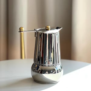 Peut inclure: Une cafetière italienne en acier inoxydable poli avec un corps cylindrique effilé, une base arrondie et un couvercle à charnière. La cafetière est ornée d'un petit bouton sphérique doré sur le couvercle et d'une boule dorée assortie sur la poignée.