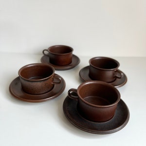 May include: Four brown ceramic teacups and saucers. The cups have small handles and sit on matching round saucers. The set is a solid, dark brown colour, and the cups and saucers are arranged on a white surface.