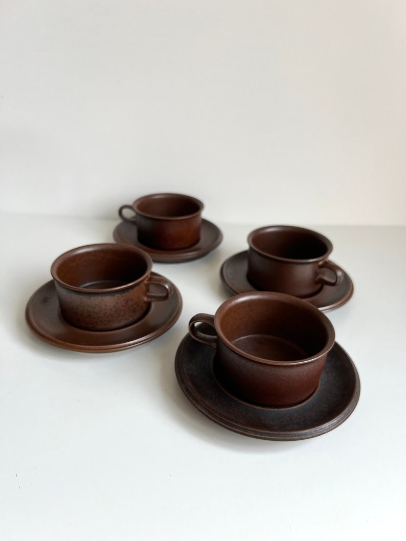 May include: Four brown ceramic teacups and saucers. The cups have small handles and sit on matching round saucers. The set is a solid, dark brown colour, and the cups and saucers are arranged on a white surface.