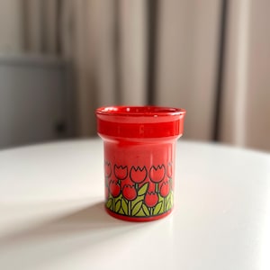 May include: Red ceramic egg cup with a tulip pattern. The cup has a white background with red tulips and green leaves.