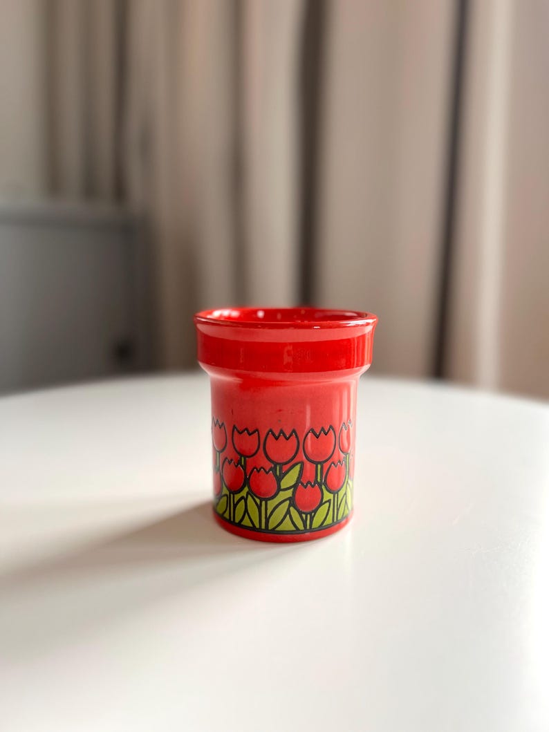 May include: Red ceramic egg cup with a tulip pattern. The cup has a white background with red tulips and green leaves.