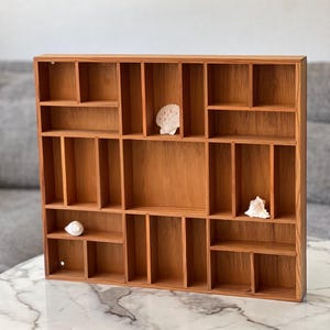 May include: A rectangular wooden display shelf featuring multiple compartments of varying sizes arranged in a grid pattern. The light brown wood grain is visible throughout the structure. Three small white seashells are placed inside the cubbies.