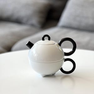 May include: White ceramic teapot with a round body and black handles. The teapot has a lid and a spout. The teapot is sitting on a white surface.