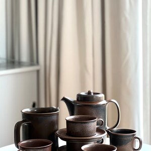 May include: A brown ceramic tea set comprising a teapot, creamer, and cups with saucers. The set has a speckled, rustic finish. The items are arranged on a white surface, with a neutral-toned curtain in the background.