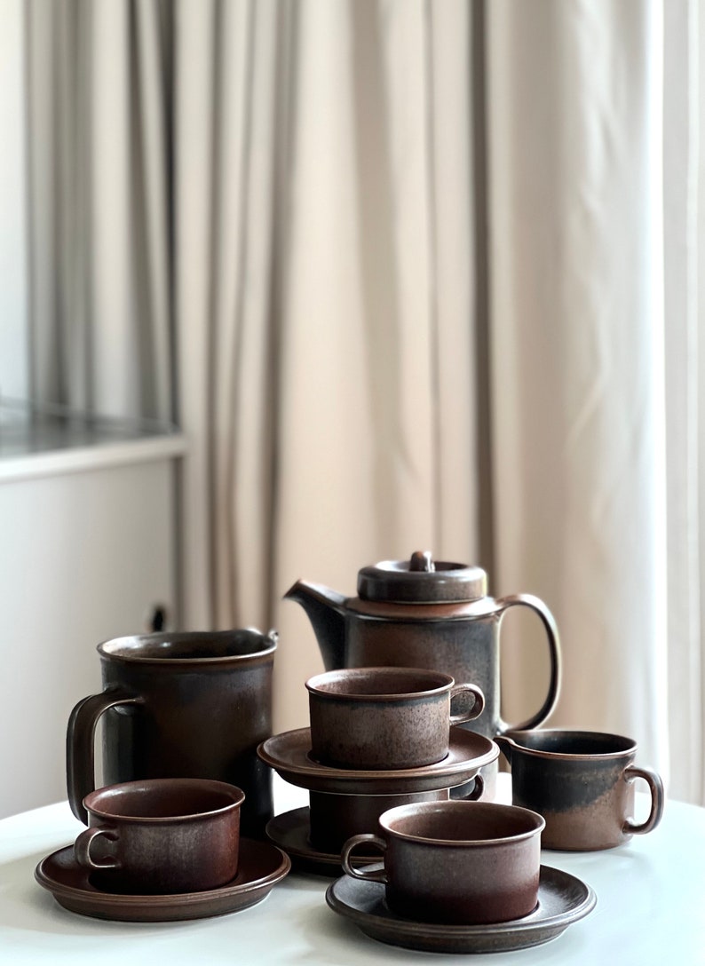 May include: A brown ceramic tea set comprising a teapot, creamer, and cups with saucers. The set has a speckled, rustic finish. The items are arranged on a white surface, with a neutral-toned curtain in the background.