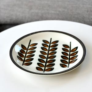 May include: A round ceramic saucer with a cream-coloured base featuring three vertical brown leaf-patterned stems. The rim of the saucer is dark brown with a textured, ribbed edge detail. The design is symmetrical, with the leaves painted in shades of brown.