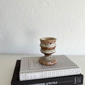 Pu&ograve; includere: Una piccola tazza in ceramica bianca con macchie marroni &egrave; appoggiata su due libri. Il libro superiore ha una copertina bianca con un motivo geometrico ripetitivo e il testo "Little Women" sul dorso. Il libro inferiore ha una copertina nera con il testo "The Handbook of Style" sul dorso.