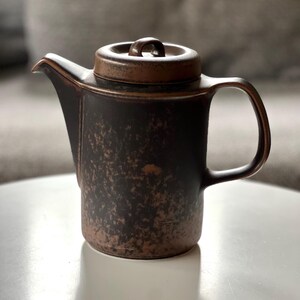 May include: A small, dark brown ceramic teapot with a matching lid. The teapot has a spout, a handle, and a textured surface. The lid has a small loop for easy removal. The teapot is likely used for brewing tea.