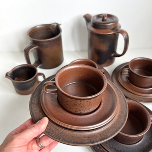 May include: A set of brown ceramic tableware, including cups, saucers, a creamer, a teapot, and a mug. The pieces have a textured surface and a warm, earthy tone. The set is arranged on a white surface.