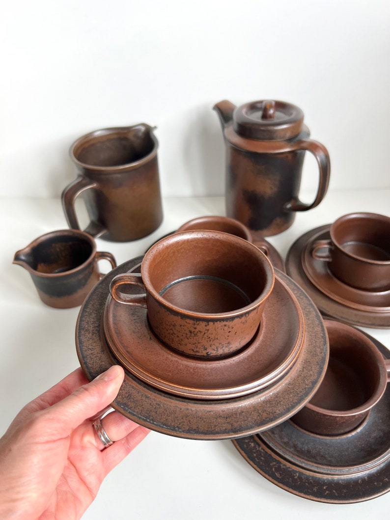 May include: A set of brown ceramic tableware, including cups, saucers, a creamer, a teapot, and a mug. The pieces have a textured surface and a warm, earthy tone. The set is arranged on a white surface.