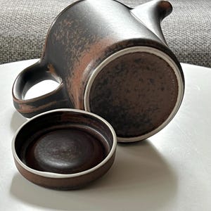 May include: A brown ceramic teapot with a matching lid. The teapot features a curved spout and a handle. The exterior has a textured, mottled appearance with shades of brown and reddish-brown. The lid is a small, round dish with a white rim.