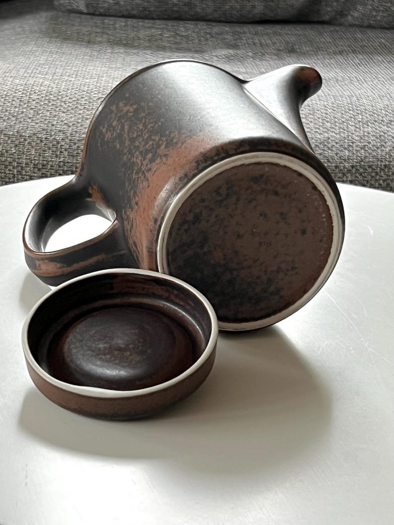 May include: A brown ceramic teapot with a matching lid. The teapot features a curved spout and a handle. The exterior has a textured, mottled appearance with shades of brown and reddish-brown. The lid is a small, round dish with a white rim.