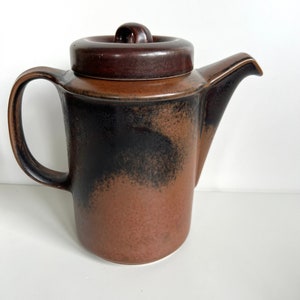May include: A brown ceramic teapot with a lid and handle. The teapot has a dark brown lid and handle, with a gradient of black to brown on the body. The spout is on the right side.