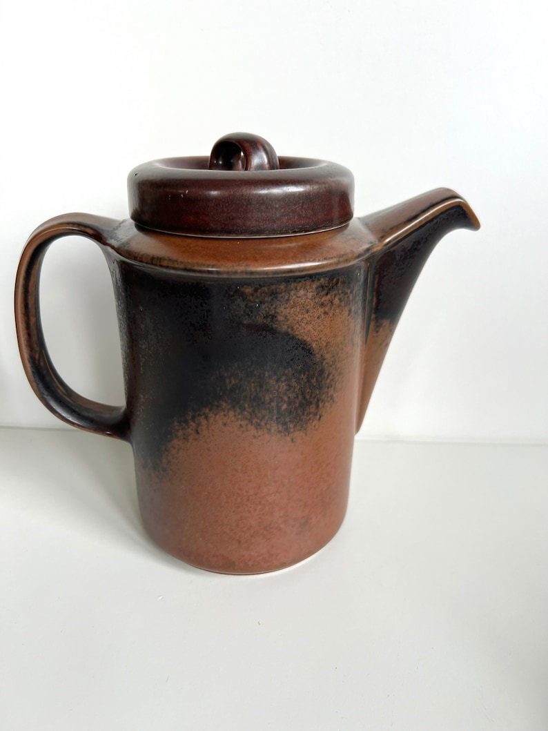 May include: A brown ceramic teapot with a lid and handle. The teapot has a dark brown lid and handle, with a gradient of black to brown on the body. The spout is on the right side.
