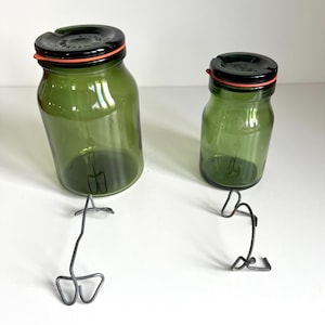 Op de afbeelding: Twee groene glazen potten met zwarte deksels en rode rubberen afdichtingen. De potten staan op een wit oppervlak met metalen potklemmen ernaast.