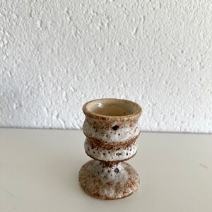 Pu&ograve; includere: Un piccolo porta uova in ceramica bianca con macchie marroni e un design testurizzato. La tazza ha una base larga e una parte superiore stretta.