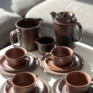 May include: A brown ceramic coffee set including a teapot, creamer, sugar bowl, and several cups and saucers. The set has a rustic, textured appearance and is arranged on a white surface. The cups and saucers are stacked.