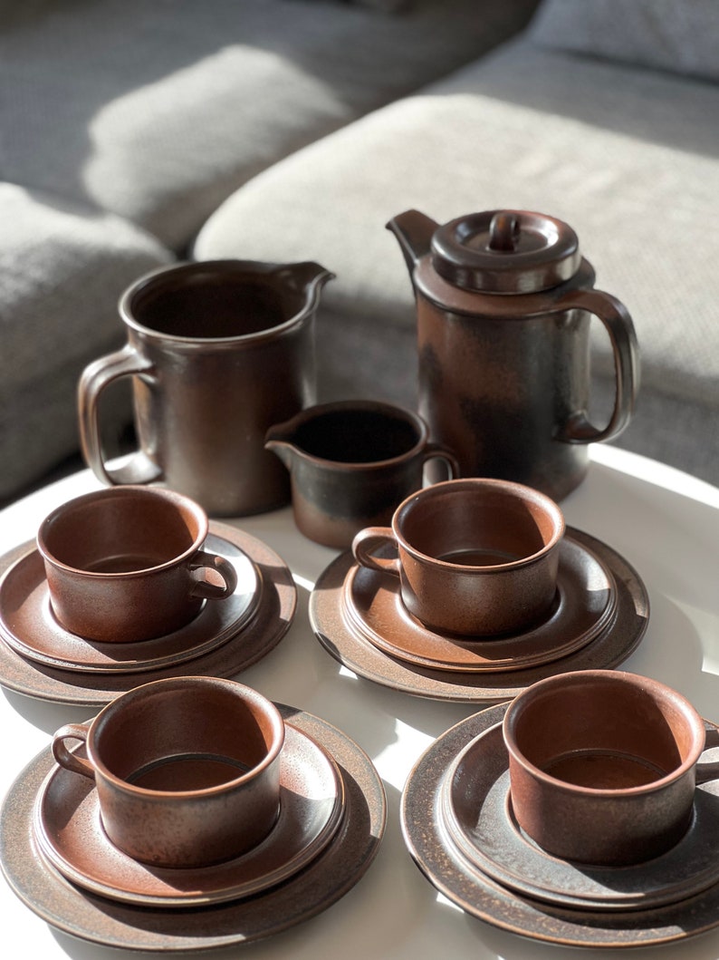 May include: A brown ceramic coffee set including a teapot, creamer, sugar bowl, and several cups and saucers. The set has a rustic, textured appearance and is arranged on a white surface. The cups and saucers are stacked.