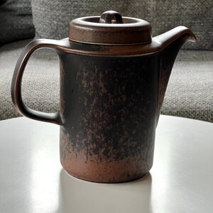 May include: A brown and black ceramic teapot with a lid and handle. The teapot has a textured surface and a spout for pouring. The handle is curved, and the lid has a small knob. The teapot is sitting on a white surface.