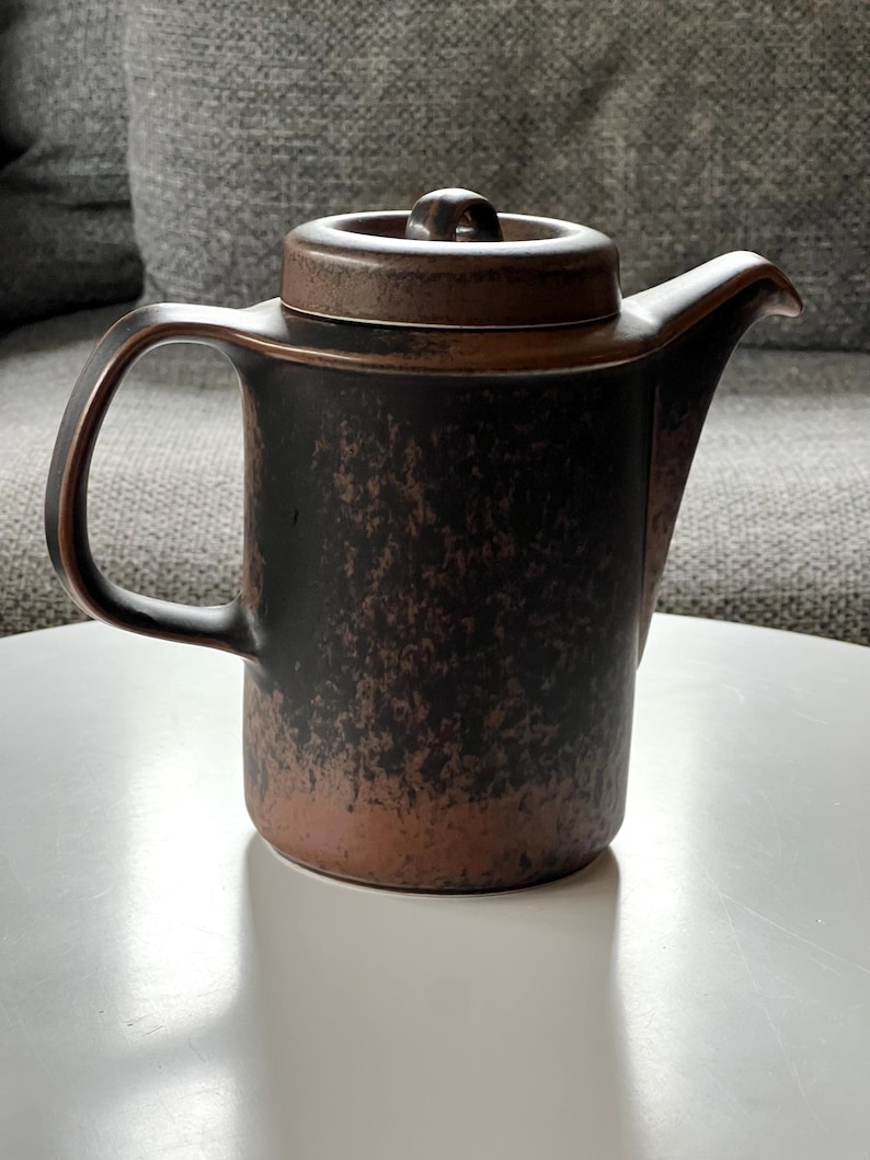 May include: A brown and black ceramic teapot with a lid and handle. The teapot has a textured surface and a spout for pouring. The handle is curved, and the lid has a small knob. The teapot is sitting on a white surface.