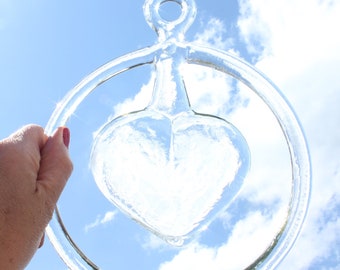 May include: Clear glass heart-shaped ornament with a circular handle. The heart is suspended within a clear ring, held against a blue sky with fluffy white clouds. The ornament is held by a hand.
