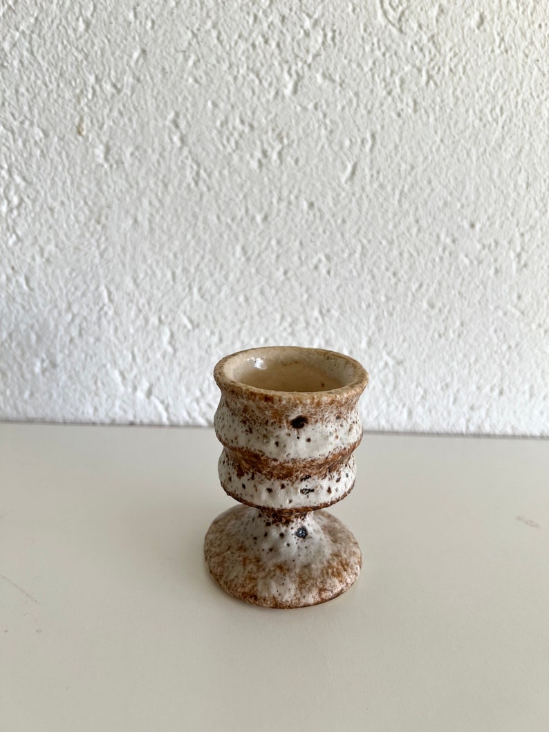 Pu&ograve; includere: Un piccolo porta uova in ceramica bianca con macchie marroni e un design testurizzato. La tazza ha una base larga e una parte superiore stretta.