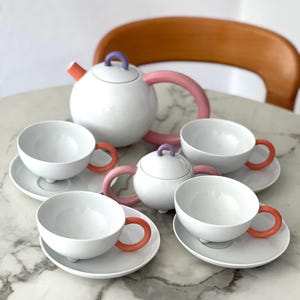May include: A white ceramic tea set with a teapot, sugar bowl, and four teacups. The teapot has a pink handle and orange spout. The teacups have orange handles. The set is on a white marble table.
