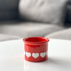 May include: A small, red ceramic pot with white heart designs around the middle. The pot is sitting on a white surface.